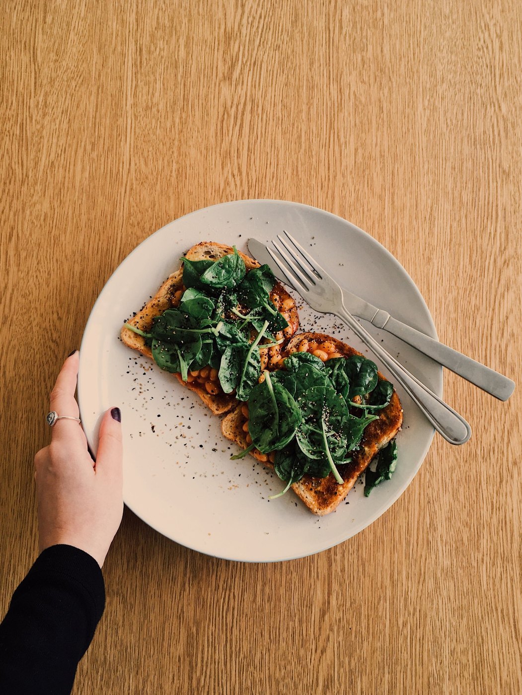 Beans on toast topped with fresh spinach on a white plate, a hand holding the plate on a wooden table