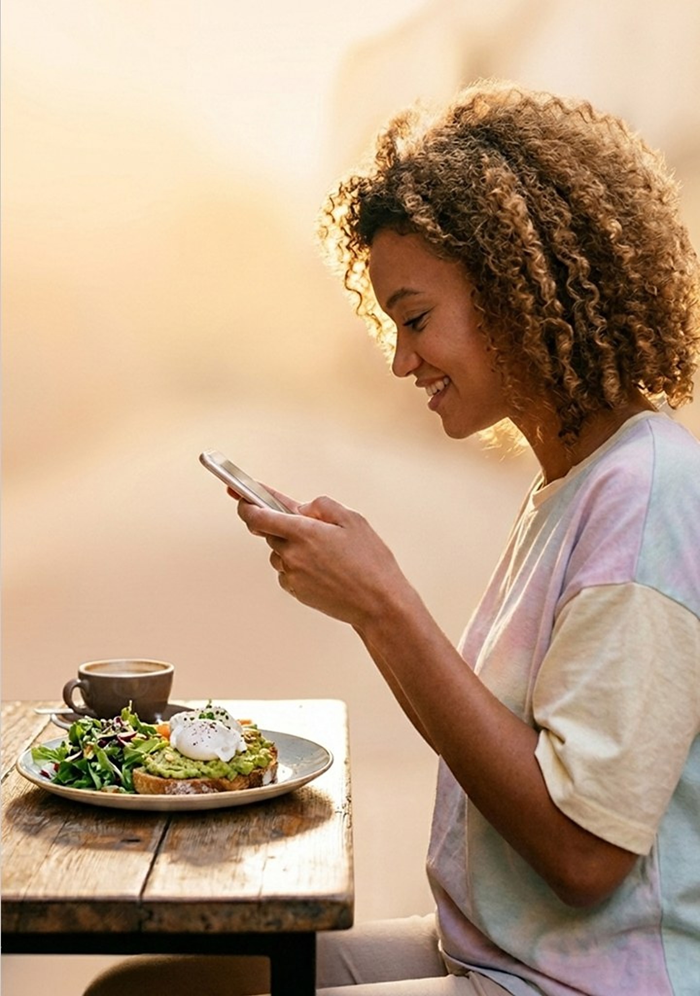 A woman smiling at her phone at a café table, with avocado toast, salad and a coffee in front of her