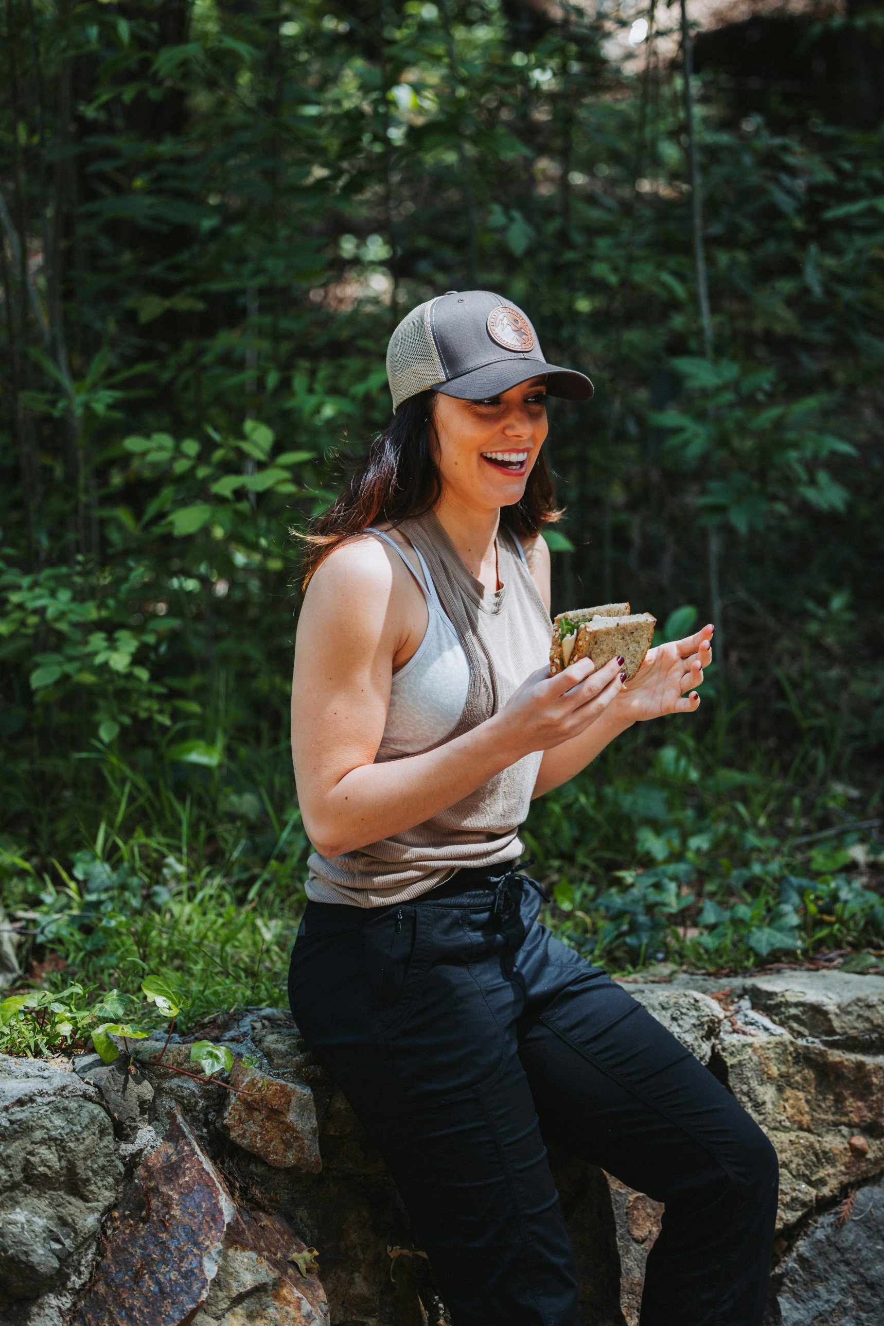 A woman in a cap laughing as she eats a wholegrain sandwich outdoors